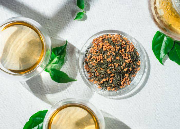 A top-down shot of genmaicha tea leaves and glass cups filled with tea. Around then, fresh green leaves.